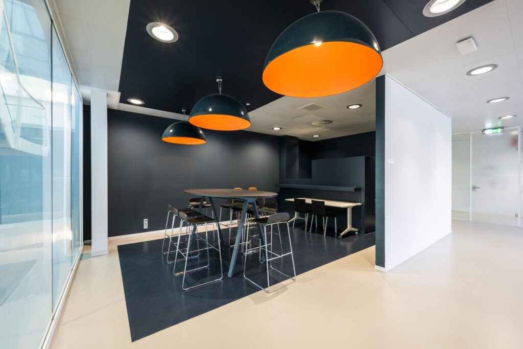 Modern meeting area with high tables, bar stools, and large dome-shaped orange pendant lights in Prof. E.M. Meijerslaan building.